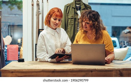 Clothing Store Partners Working On A Laptop And Digital Tablet. Two Fashion Shop Owners Standing At The Front Counter Of Their Store And Working.