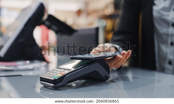 Clothing Store: Woman At Counter Buys Clothes
Paying with Smartphone Through, Contactless NFC Terminal.
Department Store, Shopping Center, Mall Purchase. Close-up Focus on
Mobile Phone