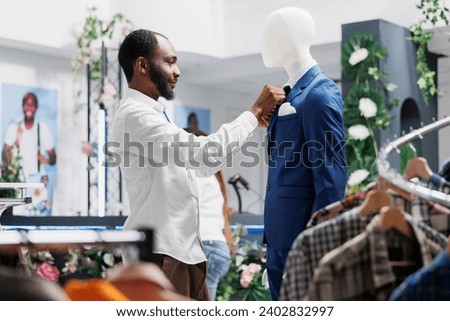 Clothing store worker fixing tie on mannequin wearing formal suit, showcasing menswear to customers. Fashion boutique african american man adjusting male trendy jacket on display dummy model