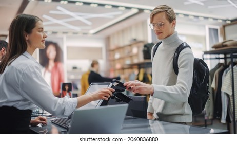 Clothing Store: Young Man At Checkout Counter Buys Clothes Paying With Contactless NFC Smartphone, Friendly Retail Sales Assistan Packs Merchandise. Contemporary Fashion Shop With Of Designer Brand