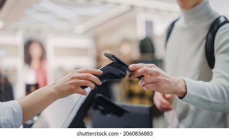 Clothing Store: Young Man At Checkout Counter Buys Clothes Paying With Contactless NFC Smartphone, Focus Of Mobile Phone And Terminal. Stylish Fashion Shop With Of Designer Brand In Big Shopping Mall