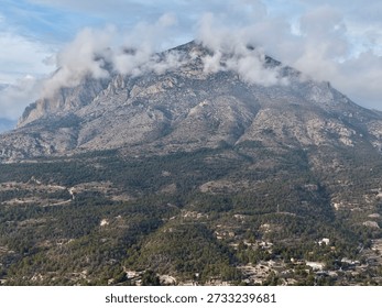 cloudcapped summit looming over forested slopes and scattered dwellings, dramatic sky and textured rock faces