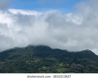 Clouds cover the peak of a mountain while the valley below shows green trees and grass. Sky is bright and clear.
