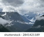Clouds envelop the mountains in a dramatic valley near Stryn, Vestland, Norway. Peaks partially obscured by mist showcase rugged terrain with patches of snow on higher altitudes. The lush green valley below contrasts with the darker, rocky mountain surfaces. A winding river or road is faintly visible, leading deeper into the misty landscape. The scene captures the moody and atmospheric essence of Scandinavian natural beauty, typical for the region during summer.