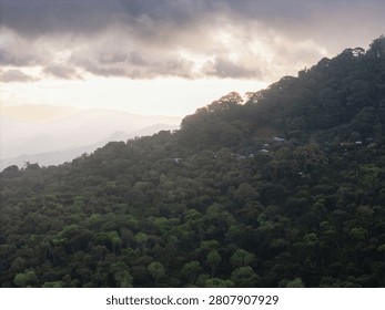 Clouds hang over a mountain as the sun sets. A small village is visible among the trees. The scene shows nature and a secluded area.