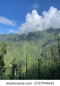Clouds hover over green mountains filled with trees. A small waterfall is visible in the distance. The scene shows nature in its usual state.