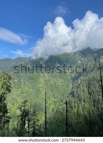 Clouds hover over green mountains filled with trees. A small waterfall is visible in the distance. The scene shows nature in its usual state.
