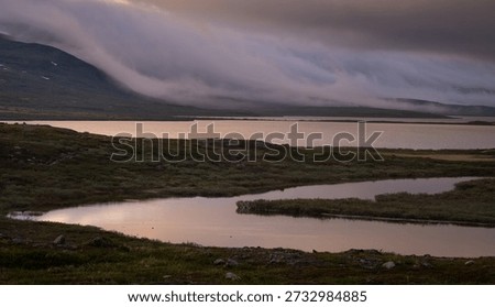 Clouds rolling down the mountains to meet Arctic lake in very low warm sunlight on Kungsleden trail. Low clouds spill down mountain slopes towards a calm Arctic lake in warm, very low polar day sunlight along the Kungsleden trail in Swedish Lapland. Soft reflections on the water and layered clouds create a dramatic, atmospheric northern evening scene.