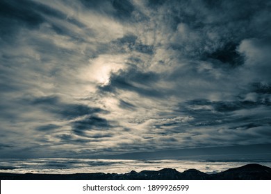Cloudscape of high clouds of various types over mountains. Sun is barely visible from the clouds in the centre. The lower level of clouds in the background behind the black mountain ridge. - Powered by Shutterstock - Get 15% off with code: PIKWIZARD15