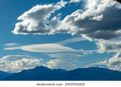 Cloudscape in mountainous area with clouds hanging above mountain peaks and large white clouds that look like waves or paint brush strokes with bright sun above