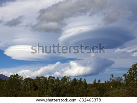 Cloudy blue sky with white clouds in the form of lenses (lenticular); Below is a green forest with coniferous trees