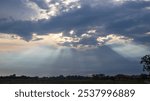 Cloudy sky with sun rays breaking through, illuminating the rural landscape below. The serene evening view creates a peaceful and atmospheric scene, perfect for themes of nature, tranquility, and coun