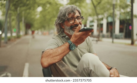 Clouse-up, smiling middle-aged man with gray hair and beard sits on bench and uses mobile phone. Mature gentleman in eyeglasses recording voice recognition message on speakerphone - Powered by Shutterstock