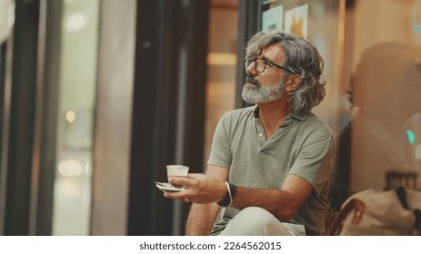 Clouse-up, thoughtful middle-aged man with gray hair and beard, wearing casual clothes, sits in street cafe. Mature gentleman in eyeglasses drinks aroma coffee - Powered by Shutterstock