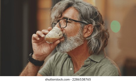 Clouse-up, thoughtful middle-aged man with gray hair and beard, wearing casual clothes, sits in street cafe. Mature gentleman in eyeglasses drinks aroma coffee - Powered by Shutterstock