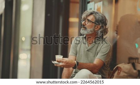 Clouse-up, thoughtful middle-aged man with gray hair and beard, wearing casual clothes, sits in street cafe. Mature gentleman in eyeglasses drinks aroma coffee