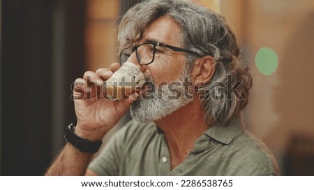 Clouse-up, thoughtful middle-aged man with gray hair and beard, wearing casual clothes, sits in street cafe. Mature gentleman in eyeglasses drinks aroma coffee