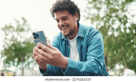 Clouseup, young bearded man in denim shirt sitting in wireless headphones making video call on mobile phone on busy street modern city background. - Powered by Shutterstock