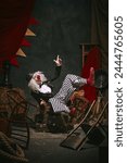 Clown in black hat, white face with red nose and striped pants playing with emotive face over dark retro circus backstage background. Concept of circus, theater, performance, show, retro and vintage
