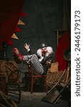 Clown in black hat, white face with red nose and striped pants performing over dark retro circus backstage background. Emotional play. Concept of circus, theater, performance, show, retro and vintage