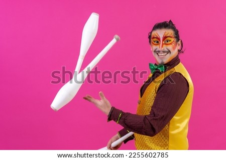 Clown with white facial makeup on a pink background, smiling juggling clubs and looking at camera