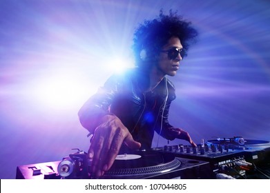 Club DJ with afro hairstyle playing mixing music on vinyl turntable at party wearing sunglasses with lens flare from nightlife lights.