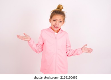 Clueless caucasian little kid girl with bun hairstyle wearing pink tracksuit over white background shrugs shoulders with hesitation, faces doubtful situation, spreads palms, Hard decision