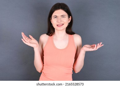 Clueless Caucasian woman wearing orange sportswear over studio grey wall shrugs shoulders with hesitation, faces doubtful situation, spreads palms, Hard decision