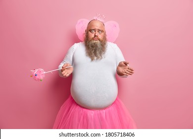 Clueless doubtful male failry wonders why his abilities disappeared, plays in performance for children, wears special costume, holds magic wand, isolated on pink background, lives at magic land