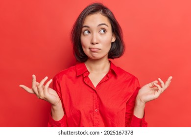 Clueless hesitant beautiful Asian woman with short dark hair spreads palms sideways shrugs shoulders has indecisive expression dressed in shirt stands against vivid red background. Who knows. - Shutterstock ID 2025167378