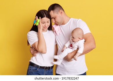 Clueless Mother And Father With Baby Posing Isolated Over Yellow Background, Couple Keep Eyes Closed, Dad Leans On Wife's Head, Holding Newborn Daughter, Mom With Beanbag Touching Head With Hand.