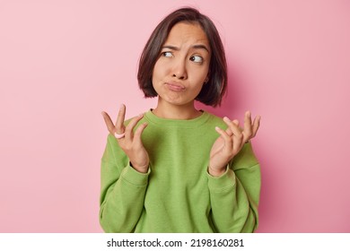 Clueless puzzled Asian woman shrugs shoulders feels hesitant about something spreads hands sideways feels unaware dressed in casual green pullover isolated over pink background. So what to do - Shutterstock ID 2198160281