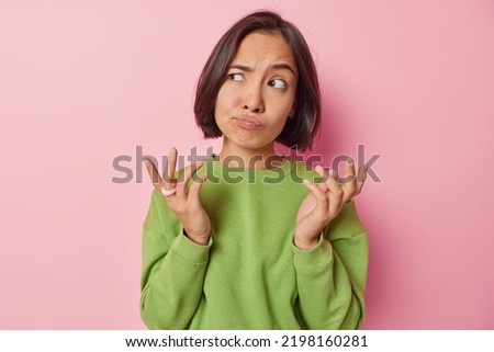 Clueless puzzled Asian woman shrugs shoulders feels hesitant about something spreads hands sideways feels unaware dressed in casual green pullover isolated over pink background. So what to do