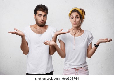 Clueless and puzzled stylish male and female standing at white wall with copy space for your text, having confused looks, shrugging shoulders as if saying: Who cares, I don't know, What's the problem