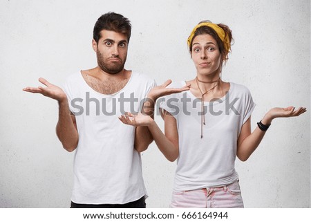 Clueless and puzzled stylish male and female standing at white wall with copy space for your text, having confused looks, shrugging shoulders as if saying: Who cares, I don't know, What's the problem