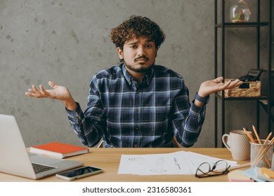 Clueless sad confused mistaken employee business Indian man he wears casual blue checkered shirt shrugging shoulders looking puzzled look camera sit work at office desk with laptop pc computer indoors - Powered by Shutterstock