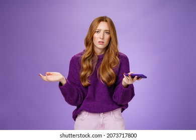 Clueless And Sad Cute Redhead Woman Spread Hands Sideways Frowning Disturbed And Upset Holding Smartphone Being Questioned And Clueless Why Mobile Phone Not Working, Posing Over Purple Wall