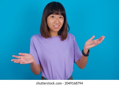 Clueless Young beautiful asian girl wearing purple t-shirt over blue background shrugs shoulders with hesitation, faces doubtful situation, spreads palms, Hard decision