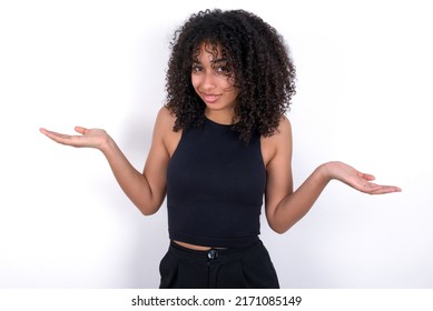 Clueless Young beautiful girl with afro hairstyle wearing black t-shirt over white background shrugs shoulders with hesitation, faces doubtful situation, spreads palms, Hard decision