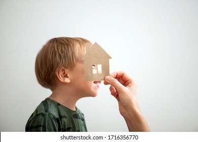 Clumsy Blond Toddler Boy In Green Long-sleeve Trying To Look Window Of Paper House Held By Mom. Over White Wall.