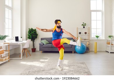 Clumsy, Funny Man In Headband And Colorful Blue, Red And Yellow Retro Sportswear Doing Front Leg Raise Toe Tap Gymnastic Exercise In Living-room. Concept Of Weight Loss And Fitness Workout At Home