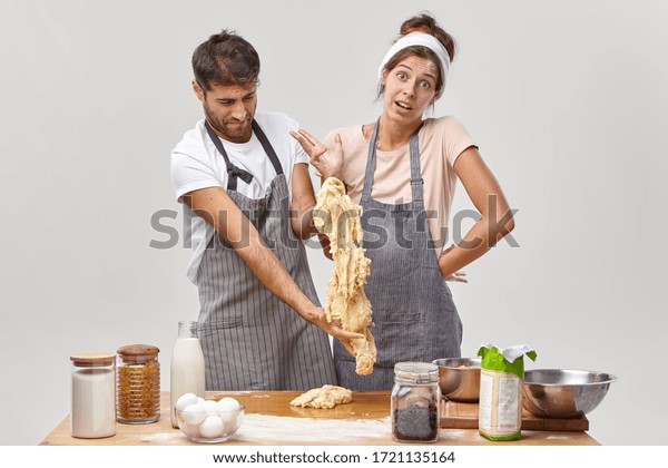 Clumsy man in apron stretches sticky dough, has\
difficulties during cooking, indignant housewife stands near,\
cannot understand whats wrong. Bad cookers at kitchen. Culinary\
courses for amateurs