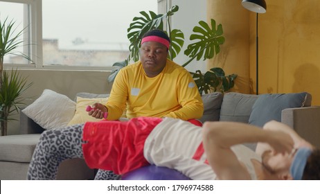 Clumsy Retro Sportsman Trying To Exercise With Fitness Ball. His Lazy Friend Black Plump Man Resting Down On Sofa Pretending To Train. Humor And Sports. Fun.