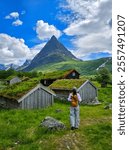 A cluster of traditional Norwegian houses with lush, green roofs nestled amidst rolling hills and towering mountains. a women visiting Innerdalen Valley Norway