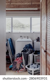 A cluttered laundry room features a washing machine surrounded by piles of dirty clothes and overflowing baskets, creating a sense of domestic disarray
