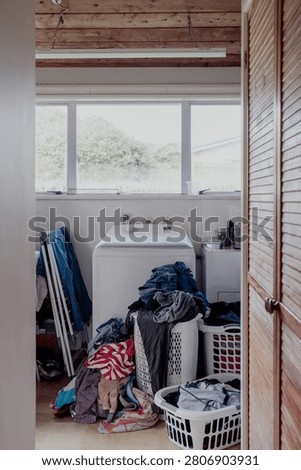 A cluttered laundry room features a washing machine surrounded by piles of dirty clothes and overflowing baskets, creating a sense of domestic disarray