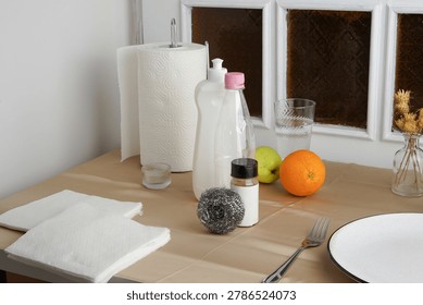 A cluttered still life of everyday objects on a beige table. The scene includes cleaning supplies, fresh fruit, paper towels, and tableware. Soft lighting highlights a mix of textures and colors