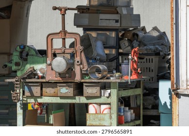 Cluttered workshop workbench with vintage hinged pipe vise and cutter surrounded by spare parts, assorted hardware, boxes, and various other materials used in heavy-duty metalwork.