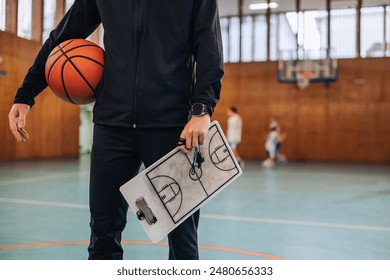 Coach in black jacket with basketball and strategy clipboard, illustrating the importance of tactics in guiding young athletes in sports - Powered by Shutterstock