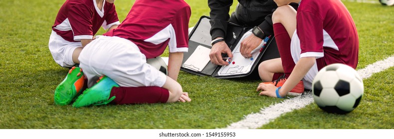 Coach Coaching Soccer Kids Soccer Team. Youth Sports Coach Using Tactics Board. Trainer Explaining Match Strategy. Sports Soccer Education. Three Junior Players Sitting With Coach On Grass Pitch